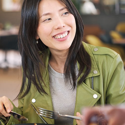 Person in green jacket seated at a café table, holding fork and knife, looking to the side.