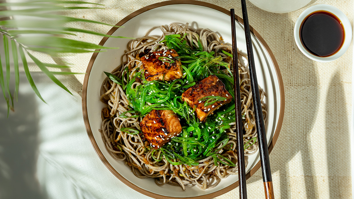 A bowl of salmon with steaming noodles garnished with green onions and served with chopsticks on the side.