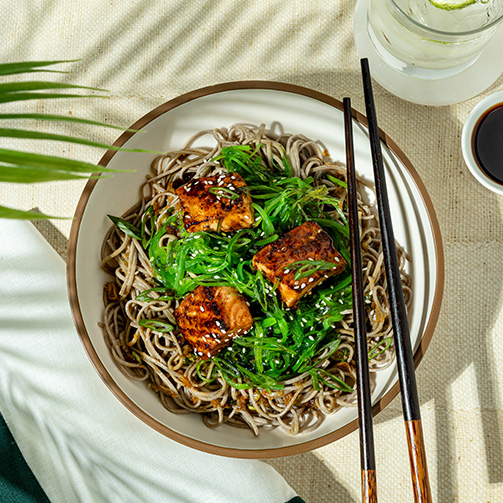 A bowl of salmon with steaming noodles garnished with green onions and served with chopsticks on the side.