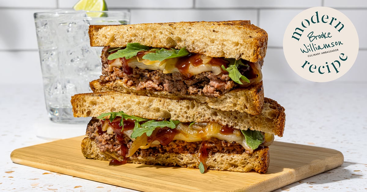 Stacked beef melt sandwiches with caramelized onions and arugula on toasted bread, on a cutting board.