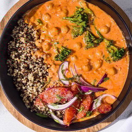 Bowl with quinoa, chickpea curry, broccoli, red onions, and blood orange slices on wood surface.