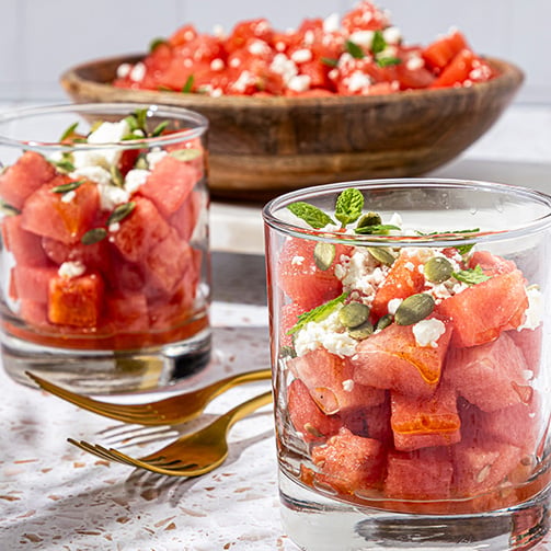 Watermelon salad in glass cups topped with feta, mint, and pumpkin seeds, with a serving bowl behind.