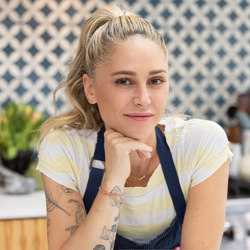 A woman with tattoos sits in a modern kitchen, surrounded by cooking utensils and a warm, inviting atmosphere.