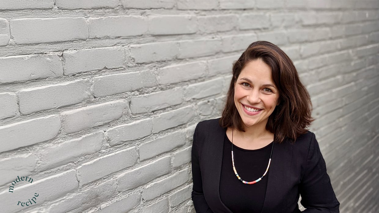 A person with shoulder-length hair stands against a light gray brick wall, wearing a black top, a blazer, and a colorful beaded necklace. A small circular logo reading "modern recipe" is visible in the bottom left corner.