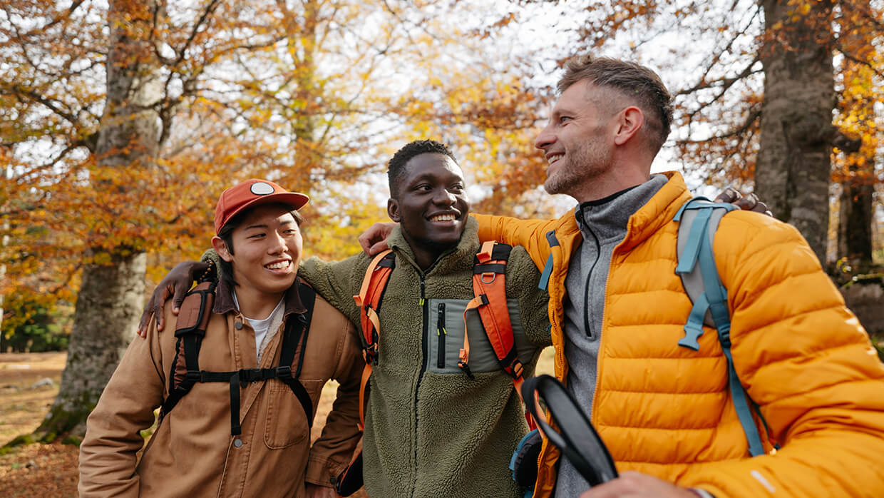 Image of three individuals outdoors with arms around each other. They are dressed in jackets and backpacks, suggesting a hiking or camping trip. The background features autumn-colored trees, emphasizing a seasonal setting and camaraderie in nature