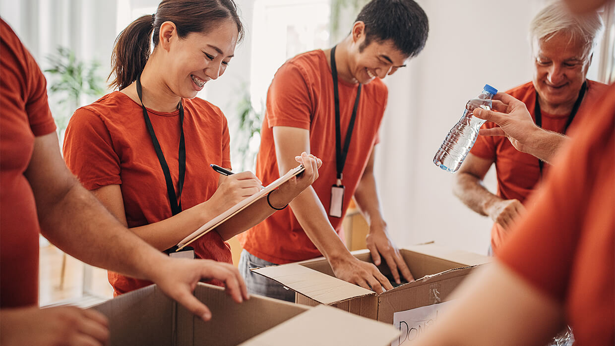 Image of a group of people wearing orange shirts and black lanyards working together on a project involving cardboard boxes labeled for donations. One person holds a clipboard and writes, while another holds a bottle of water, highlighting teamwork and community service