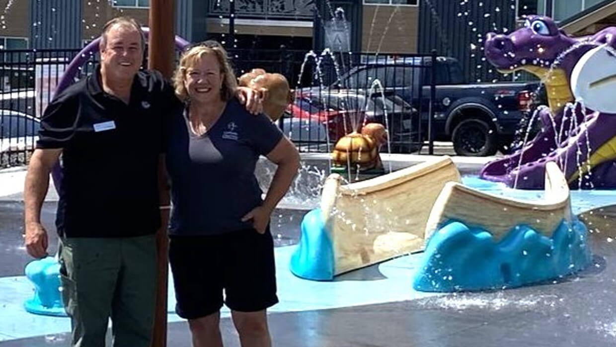 Image of two individuals standing in front of a water play area with fountains and playful structures, including a purple dragon and a boat surrounded by blue waves. Their faces are blurred, and they are posing with their arms around each other in an outdoor recreational setting