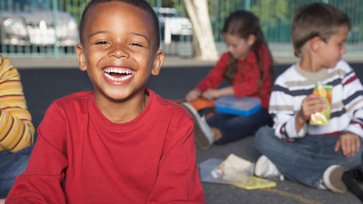 Image of three children sitting on the ground outdoors, sharing snacks and drinks. The child in the foreground wears a red shirt, while the others are dressed in striped shirts with vibrant colors. Lunchboxes and snacks are scattered around them, and a fence is visible in the background, suggesting a casual outdoor setting