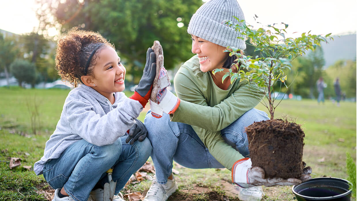 Image of an adult and a child working together to plant a small tree outdoors. The adult holds the tree with exposed roots, while the child uses a small gardening tool. Both wear gloves, emphasizing teamwork and environmental conservation