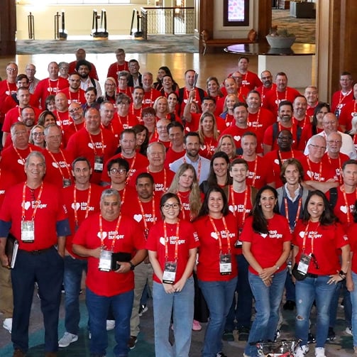 Large group in red shirts with white text and name badges gathered in a lobby or conference area with columns, railings, planters, and tables in the background.