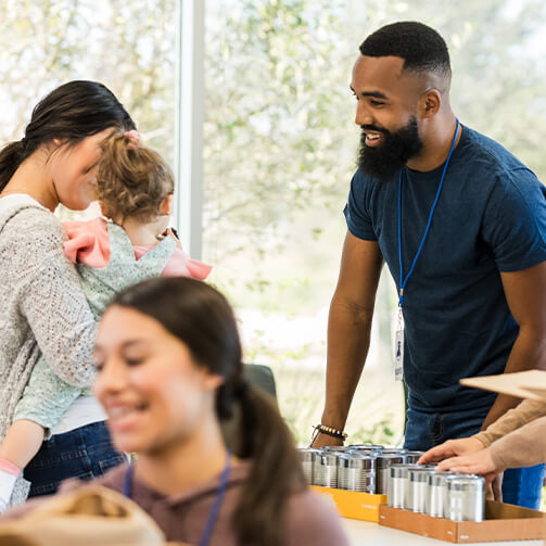 A volunteer with a beard and a lanyard smiles while assisting a mother holding a young child at a donation table. Other volunteers work in the background.