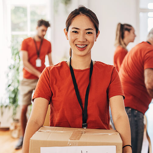 Image showing a group of individuals dressed in red shirts and black lanyards engaged in a community service activity. The person in the foreground is holding a cardboard box, suggesting involvement in organizing or distributing items. The teamwork and collaborative spirit of the group are highlighted in the indoor setting, likely a community center or similar space
