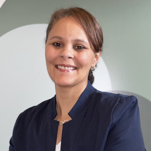Person wearing a dark blazer standing in front of a light-colored geometric background.