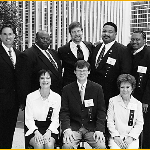 Eight formally dressed individuals with name tags, arranged in two rows (five standing, three seated) against an urban backdrop of tall buildings. Faces are blurred for privacy