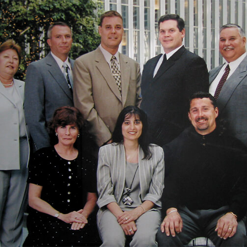 Seven formally dressed individuals (four men, three women) pose in a group photo, with some standing and others seated against a building with vertical lines and greenery.