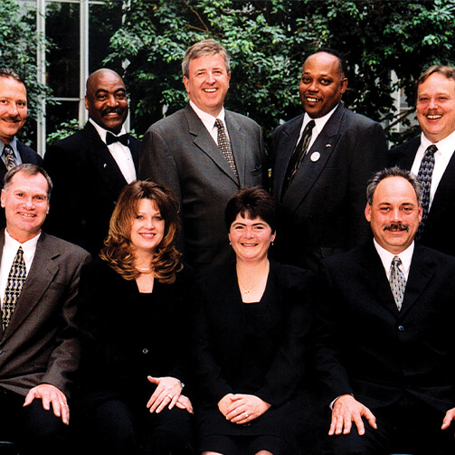 Nine individuals in formal attire with blurred faces pose indoors near greenery.
