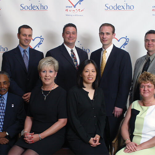Eight individuals in formal attire (five standing and three seated) at a Sodexho event, with a banner reading 'Sodexho' and 'STOP Hunger Making a Difference' alongside a heart-shaped logo