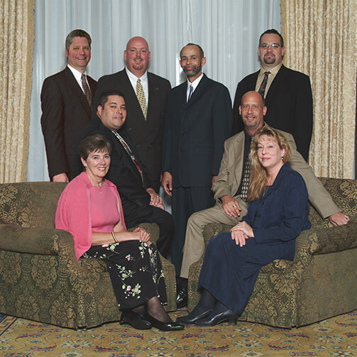 Eight individuals in formal attire, arranged with some standing and others seated on a couch against a backdrop of curtains and a patterned carpet