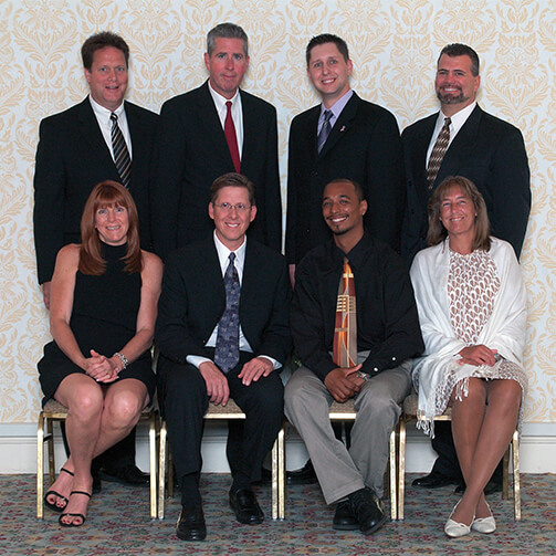 Eight individuals in formal attire: four men stand in the back row while two men and two women sit in the front row on chairs, with patterned wallpaper and a carpeted floor in the background