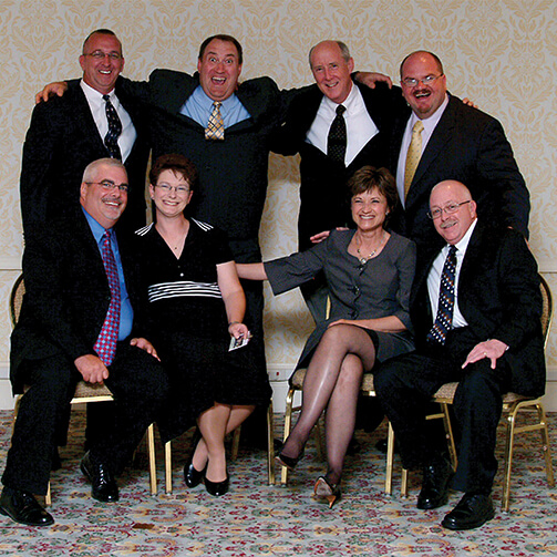 Group photo of eight people at a formal event: five men in suits stand in the back while three sit in the front—with one woman notably seated on an armrest—against a patterned wallpaper and floral carpet.