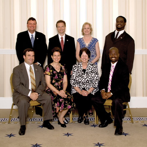 Group photo of seven individuals in formal attire. Three people are seated in the front row while four stand in the back, set against a light-striped wall with a white chair rail and a star-patterned carpet.