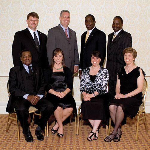 Group photo of seven individuals in formal attire: three people stand in the back while four sit in the front, set against a patterned wallpaper and carpeted floor.