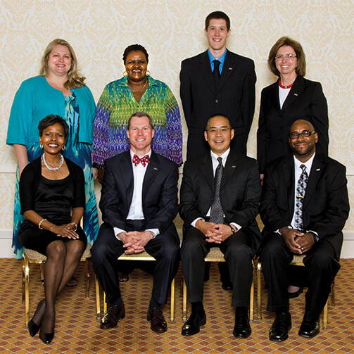 Eight individuals in formal attire pose for a group photo: four seated in the front row and four standing in the back, set against a backdrop of patterned wallpaper and a carpeted floor.