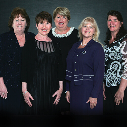 Group photo of five individuals in formal, dark-colored attire, posed against a plain dark background. Some outfits feature decorative patterns and accessories.