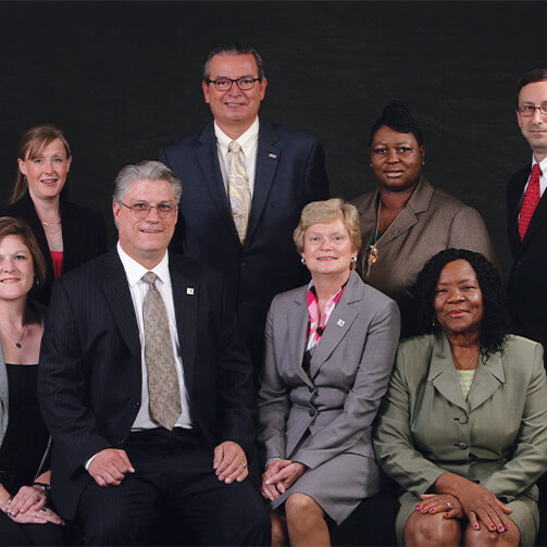 Group of twelve individuals in formal business attire posing for a professional photograph against a dark curtain, arranged in two rows on a patterned carpet.