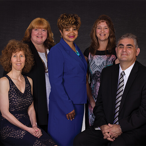 Group of five adults posing for a formal photograph, dressed in elegant attire, with a dark background emphasizing their silhouettes