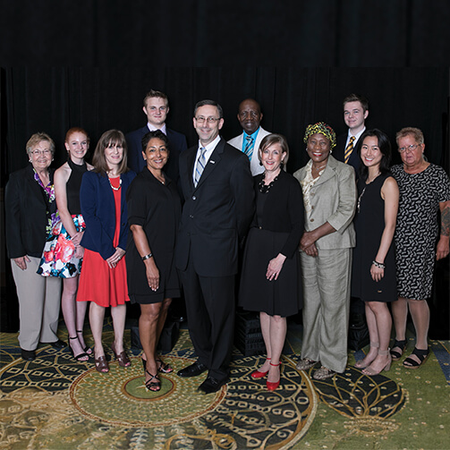 Group of twelve people standing together in formal attire in front of a dark curtain, arranged in two rows on a patterned carpet, with some leaning slightly forward.