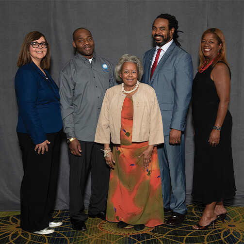 Group of five individuals standing together in front of a gray backdrop, dressed in formal and semi-formal attire.