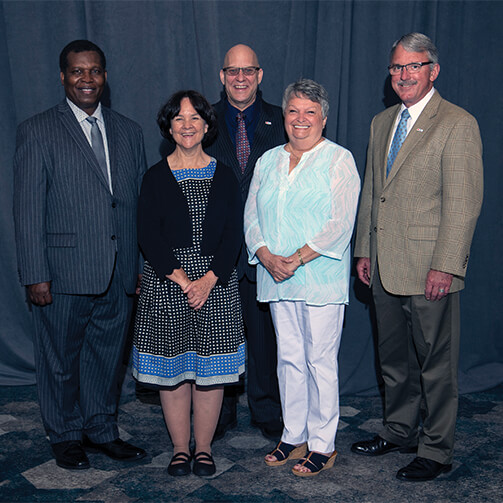 Five individuals stand together in a formal setting, dressed in professional and semi-formal attire. They are positioned in front of a plain dark curtain and on a patterned carpet