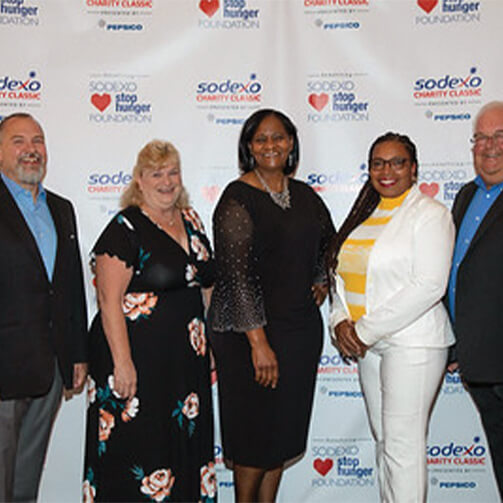 Five individuals stand in front of a backdrop displaying logos and text for the Sodexo Stop Hunger Foundation and Sodexo Charity Classic, along with the PepsiCo logo. They are dressed in formal attire, and their faces are blurred for privacy
