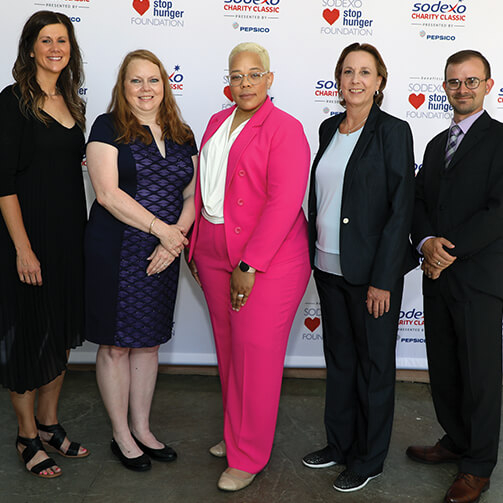 Five individuals in business or semi-formal attire stand before a backdrop featuring the logos for the 'Sodexo Stop Hunger Foundation,' 'Sodexo Charity Classic,' and 'PepsiCo.' The setting indicates a charity event focused on combating hunger