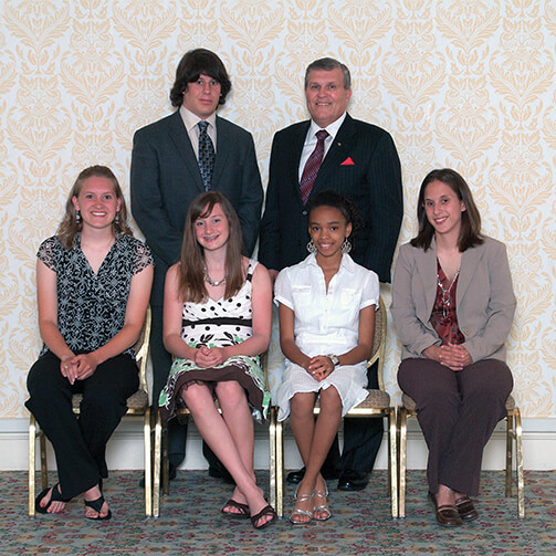 Group photo of six individuals in formal attire: two men stand in the back row while four women sit in the front on chairs, set against a patterned wallpaper backdrop.