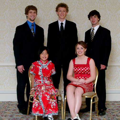 Group photo of five individuals in formal attire: three standing in suits and ties, and two seated in red dresses—one with a floral pattern and one with white polka dots—set against patterned wallpaper and carpet.