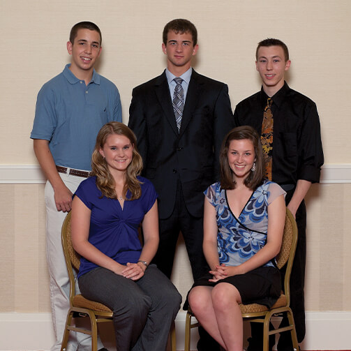 Five individuals pose for a formal photograph in front of a light-colored wall with a white chair rail, with two seated in the front and three standing behind, dressed in a mix of business and casual attire
