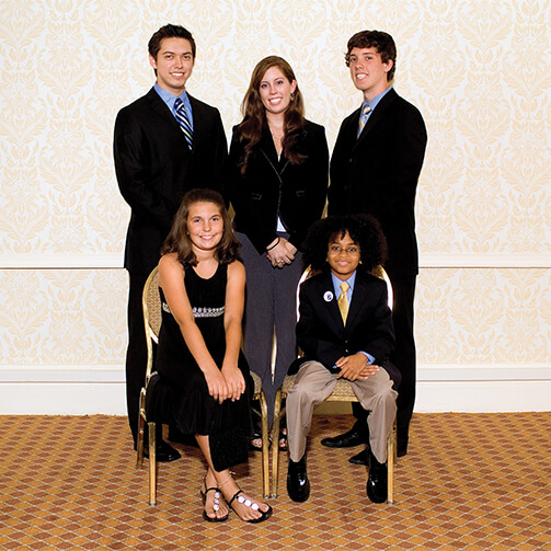 Group of five individuals posing for a formal photograph in front of patterned wallpaper, with two seated in the front row and three standing behind, dressed in suits and a dress, on a patterned carpet.