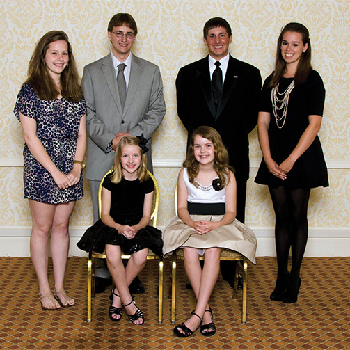 Group photo of six individuals in formal attire: four standing and two seated on chairs, set against patterned wallpaper and a carpeted floor