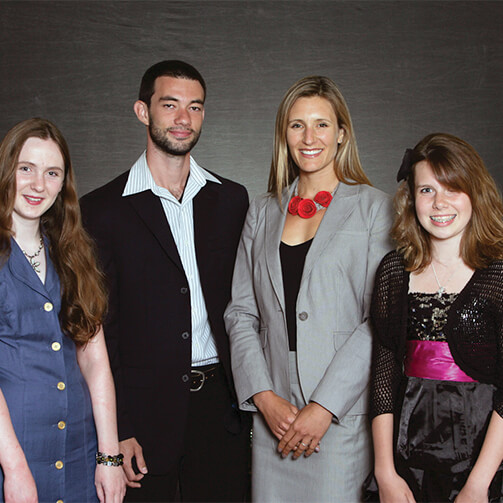 Four individuals stand side by side against a dark background in formal attire: one in a blue dress, one in a dark suit, one in a light gray suit with a red necklace, and one in a black dress with a pink sash.
