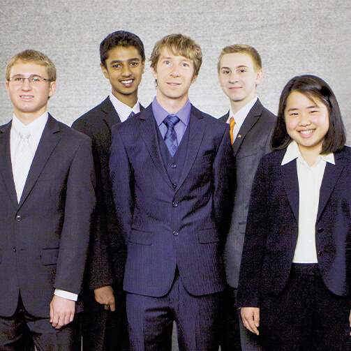 Five individuals in formal business attire stand in a row against a plain, light-colored wall, with three in the front and two behind.