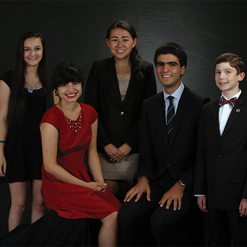 Group photo of five individuals in formal attire: three standing in the back row and two seated in the front, with a dark background contrasting their elegant clothing.