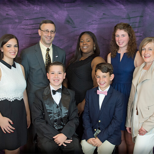 Group photo of seven individuals in formal attire, posed against a textured purple backdrop. The group includes men in suits and bow ties and women in elegant dresses, suggesting a formal event or gathering