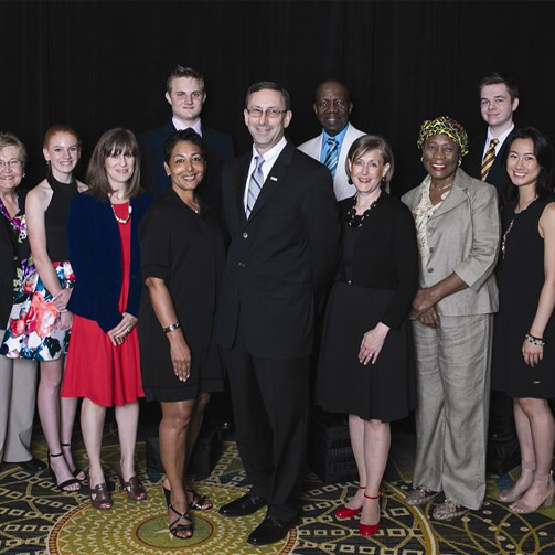 A group of eleven individuals dressed in business and semi-formal attire stands together in a professional setting. The background is dark, and the group is positioned on a patterned carpet, suggesting a formal event or gathering.