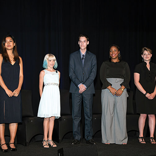 Five individuals stand in a row on a stage, dressed in formal attire. Three wear dresses while two wear suits. Chairs are visible in the background against a dark setting.