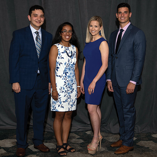 Four individuals stand side by side, dressed in formal attire. Two are in suits, while the other two wear dresses. The background features a dark curtain, and the floor has a patterned design, suggesting a formal event or gathering