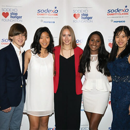 Five individuals stand in front of a backdrop featuring logos and text for the 'Sodexo Stop Hunger Foundation' and 'Sodexo Charity Classic' presented by PepsiCo. They are dressed in formal attire, indicating a charity event or gala setting