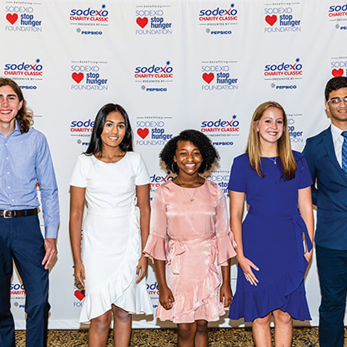 Five individuals stand in front of a backdrop featuring logos for the 'Sodexo Charity Classic,' 'Stop Hunger Foundation,' and 'PepsiCo.' Dressed in formal attire, the group appears to be at a charity event or fundraiser.