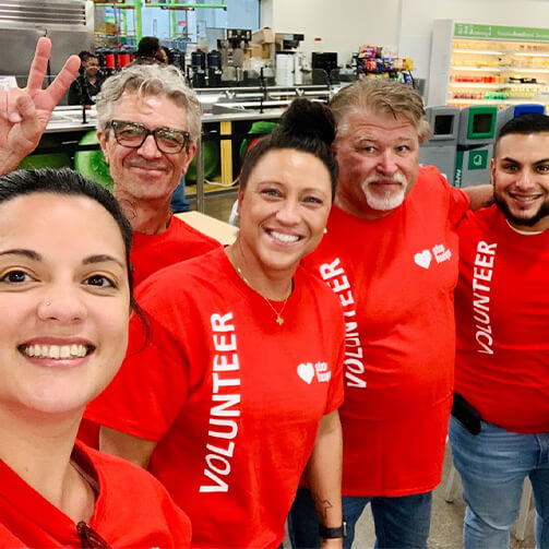 A diverse group of five cheerful volunteers in red “Volunteer” t-shirts pose for a selfie in a cafeteria, showing team spirit and enthusiasm.